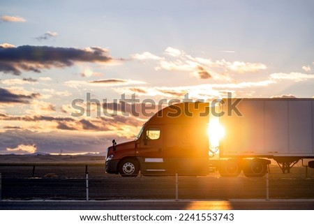 Industrial long hauler big rig red semi truck tractor with chrome parts transporting commercial cargo in loaded dry van semi trailer running on the highway road at sunset time in California Royalty-Free Stock Photo #2241553743