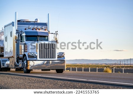Long extended cab big rig white classic powerful stylish semi truck tractor with chrome parts and satellite antenna transporting cargo on step down semi trailer running on the straight highway road Royalty-Free Stock Photo #2241553765