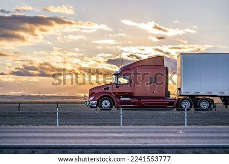 Industrial long hauler big rig burgundy semi truck tractor with chrome parts transporting commercial cargo in loaded dry van semi trailer running on the highway road at sunset time in California Royalty-Free Stock Photo #2241553777