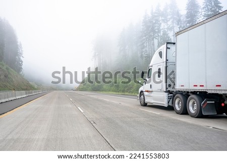 White big rig long haul semi truck transporting commercial cargo in dry van semi trailer running on turning one way highway road with forest on the hill at strong fog weather with poor visibility  Royalty-Free Stock Photo #2241553803