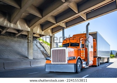Orange Big rig long haul industrial semi truck tractor with chrome accessories transporting commercial cargo in dry van semi trailer running for delivery on the summer highway road under the bridge Royalty-Free Stock Photo #2241553811