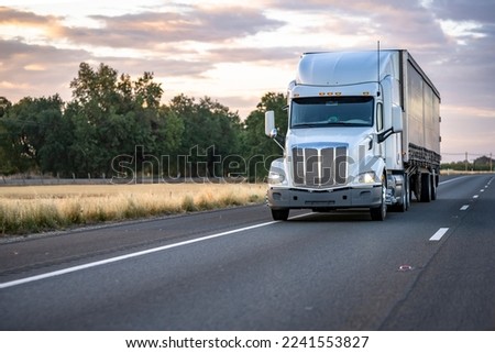 White big rig long haul industrial semi truck with turned on headlight transporting commercial cargo in tented black dry van semi trailer running on the evening highway road at twilight time Royalty-Free Stock Photo #2241553827