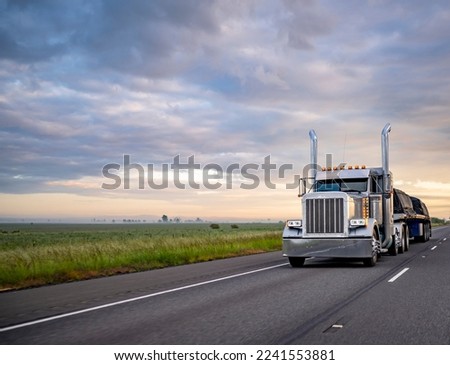 Gray classic big rig American semi truck with turned on lights and chrome exhaust pipes transporting covered cargo on flat bed semi trailer driving on the twilight highway road with stormy clouds sky Royalty-Free Stock Photo #2241553881