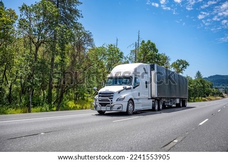 White big rig long haul day cab semi truck transporting commercial cargo in tented black dry van semi trailer with front wall spoiler running on one way highway road with trees and hill on background Royalty-Free Stock Photo #2241553905