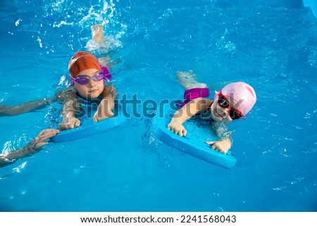 Two little girls learning how to swim in swimming pool using flutter boards