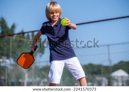 Happy blonde boy playing pickleball game, hitting pickleball yellow ball with paddle, outdoor sport leisure kids activity. Royalty-Free Stock Photo #2241570373