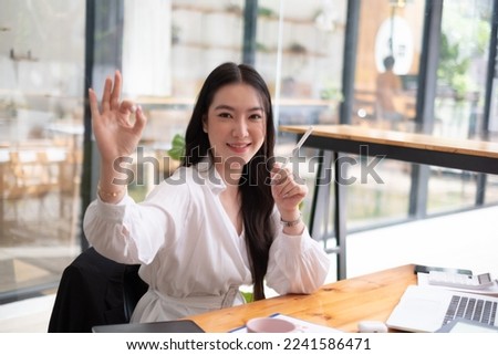 Beautiful asian businesswoman smiling and showing OK sign while sitting at her desk office, successful business concept.