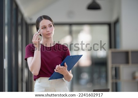Attractive businesswoman standing and thinking while holding a pen and a folder of documents.