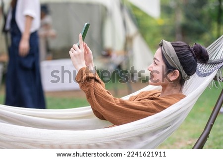 Asian woman taking a picture with a smartphone while riding a hammock at a campsite (selfie)