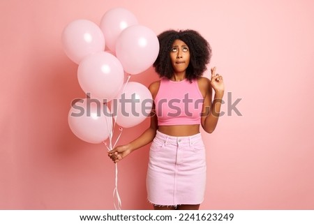 Pretty mixed race woman, makes a celebrates isolated, holds balloons, crossed her fingers in a wish and looks up, to the copy space