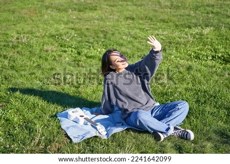 Happy asian girl sitting on picnic blanket with ukulele, cover herself from sunlight, extending hand towards sun beams and smiling.
