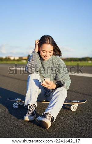 Vertical shot of asian woman sitting on skateboard on road, holding smartphone app. Skater girl skates on longboard, using mobile phone.