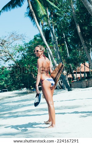 Fashion lifestyle outdoors. Sporty young woman with longboard in hand walking on white sand on tropical beach
