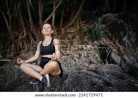 A woman in a black sports top and shorts sitting on the rocks in the tropics is engaged in sports breathing.