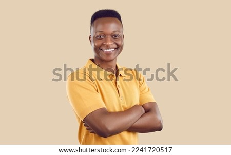 Portrait of happy young man in casual clothes. Joyful handsome well shaved African American guy in sandy yellow T shirt standing with his arms folded on beige background, looking at camera and smiling Royalty-Free Stock Photo #2241720517
