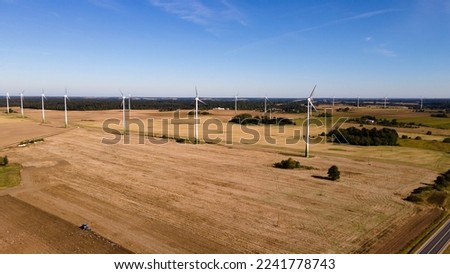 Several wind turbines stand in a row in a beautiful golden wheat field. Against the backdrop of a bright blue sky with clouds, and green trees and bushes. Photo from a bird's eye view, from a drone. S
