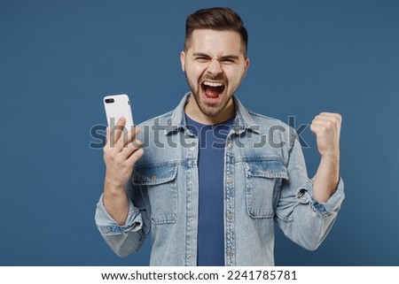 Excited young brunet man 20s wears denim jacket hold in hand use mobile cell phone talk speak on mobile cell phone celebrating clenching fists say yes isolated on dark blue background studio portrait.