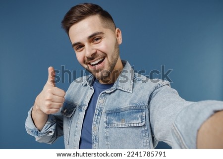 Close up happy young brunet man 20s wears denim jacket do selfie shot on mobile phone showing thumb up like gesture isolated on dark blue background studio portrait. People emotions lifestyle concept