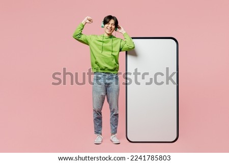 Full body fun young man of Asian ethnicity wear green hoody headphones listen to music near big huge blank screen mobile cell phone smartphone with area isolated on plain pastel light pink background