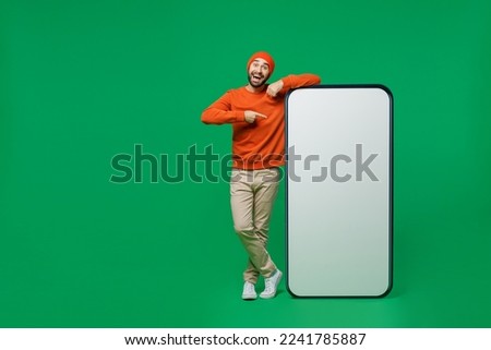 Full body young smiling fun man 20s wear orange sweatshirt hat point index finger on big blank screen mobile cell phone with workspace copy space mockup area isolated on plain green background studio