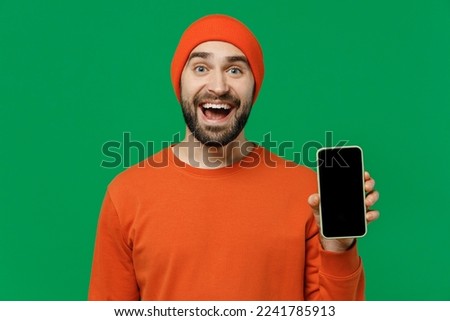 Young surprised happy fun man 20s wear orange sweatshirt hat hold in hand use mobile cell phone with blank screen workspace area isolated on plain green background studio. People lifestyle concept.