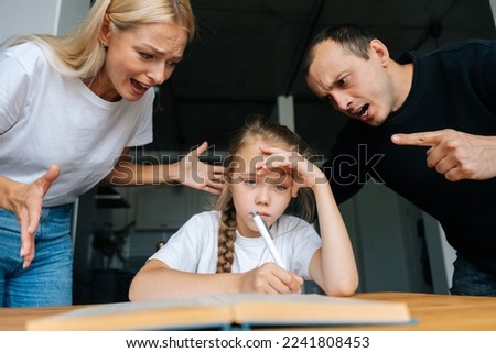 Portrait of angry young parents yelling and scolding together lazy little daughter sitting at table, doing homework, sad looking at camera. Concept of parent disciplining child for bad education. Royalty-Free Stock Photo #2241808453