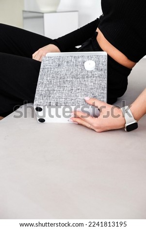 An elegant girl with a smart watch on her hand sits on a sofa and holds a bluetooth speaker in her hands. Vertical photo