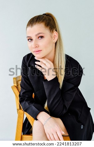 Beautiful serious girl in a black jacket sits on a chair on a gray background. Vertical photo