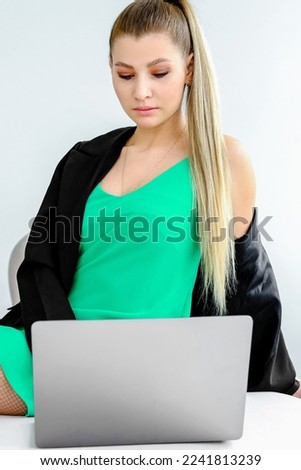 A beautiful slender fashionable girl in a green dress and a black jacket sits on a sofa and looks into a laptop. Girl buys fashion items online. Vertical photo
