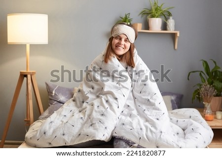 Image of satisfied delighted cheerful beautiful woman in blindfold sitting on bed wrapped in duvet, looking at camera with toothy smile and happy expression, weekend morning.