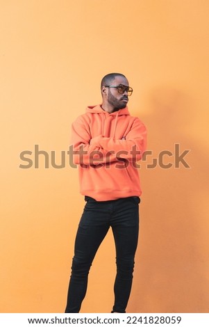 Handsome African male fashion model wearing sunglasses and seriously posing with crossed arms while looking to the side, he is standing near the orange wall outside. African man smiling over isolated 