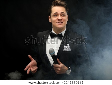 Stylish young man in a tuxedo holding a microphone, posing against a dark background with smoke, actor, singer, show, host of the event. Party concept. Royalty-Free Stock Photo #2241850961