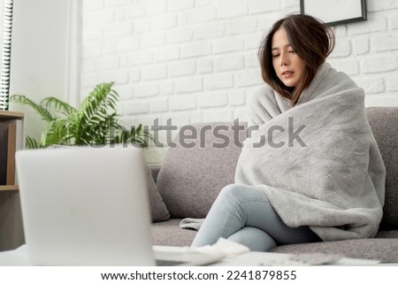 Sick Asian woman feeling cold covered with blanket sitting on sofa watching movie on laptop. Cold and fever concept. Royalty-Free Stock Photo #2241879855