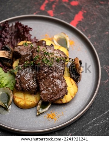 Beefsteak garnished with fried potatoes, mushrooms and lettuce on dark metal plate. Grill menu. Grilled meat. Serving the dish. View from above. Soft focus. Dark background.