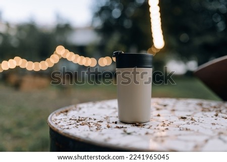 A thermos cup for hot drinks stands outside on a table against a background of warm light bulbs. Mockup or photo for advertising