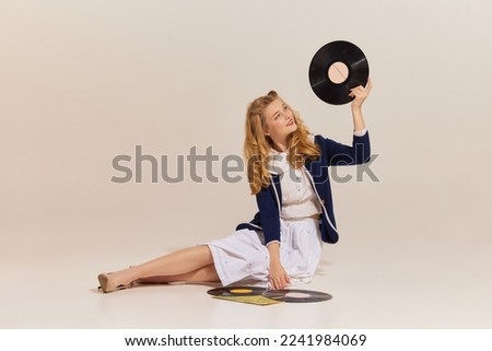 Music. Happy beautiful stylish young blonde woman with retro style hairdo wearing vintage outfit sitting on floor with vinyl record isolated over grey background. Beauty, ad, emotions Royalty-Free Stock Photo #2241984069
