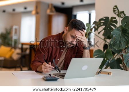 Man working on laptop while sitting at his working place in his apartment. Tired brown haired man taking glasses off working too long at computer. Exhausted male suffer from headache. Royalty-Free Stock Photo #2241985479