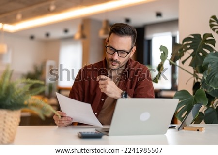 Serious millennial man using laptop sitting at the table in a home office, focused guy looking at the paper, communicating online, writing emails, distantly working or studying on computer at home. Royalty-Free Stock Photo #2241985507