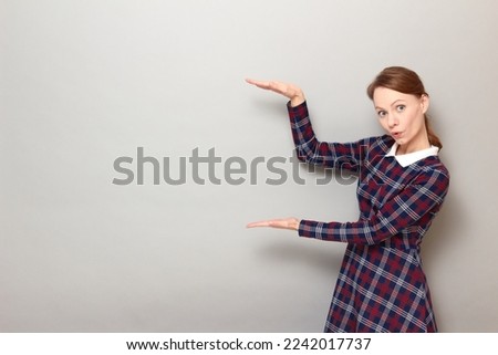 Portrait of surprised young woman showing size of something big with hands, with empty place for your text and design, looking amazed and impressed, wearing dress, standing over gray background