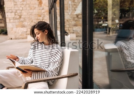 Modern young caucasian girl leafing through notebook, holding in her hands sitting on armchair street. Brunette woman with wavy hair wears casual clothes. Concept of learning.