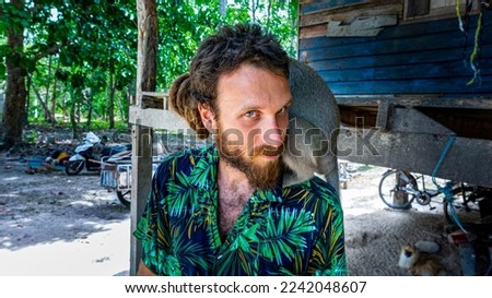 Alternative white European male person with a grey monkey on his shoulders in a rural village on a tropical Thai island on a beautiful sunny day. 