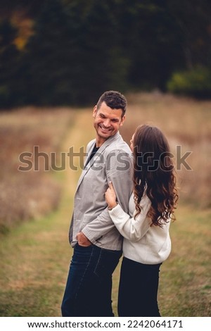 A happily married couple smiling at each other with the wife tugging at her husband's arm in a sunny field in the countryside Royalty-Free Stock Photo #2242064171