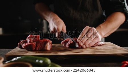 Chef sharpening his knife in front of raw piece of steak. Cooker preparing his tools for cutting meat for grilling on professional kitchen table Royalty-Free Stock Photo #2242066843
