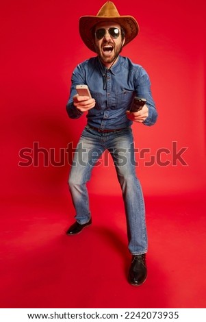 Excited young man in cowboy style outfit having fun, posing isolated over red background. Model in cowboy hat and denim clothes. Fashion, emotions concept. Looks happy, delighted