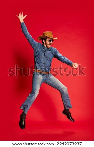 Excited young man in cowboy style outfit having fun, posing isolated over red background. Model in cowboy hat and denim clothes. Fashion, emotions concept. Looks happy, delighted