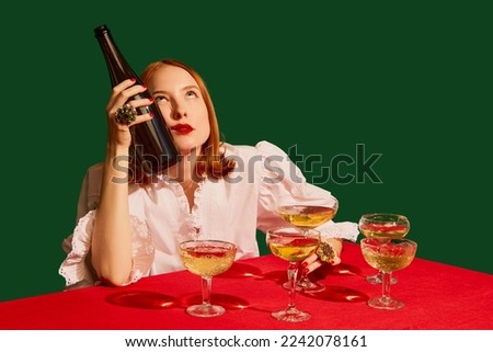Young redhead girl in festive white dress sitting at the table with champagne over green background. Hangover. Concept of holiday, party, drink. Complementary colors. Copy space for ad. Pop art