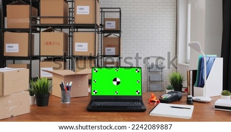 Laptop with green screen chromakey display mockup standing on desktop in warehouse. Cardboard boxes in the background.