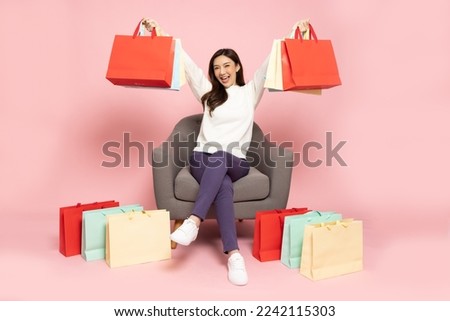 Full length portrait of Excited Asian woman sitting on armchair with shopping bags isolated on pink background, Shopper or shopaholic concept Royalty-Free Stock Photo #2242115303