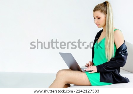 A beautiful serious girl in a green dress and a black jacket is sitting on a sofa with a laptop on her lap. girl looking at laptop screen. Horizontal photo