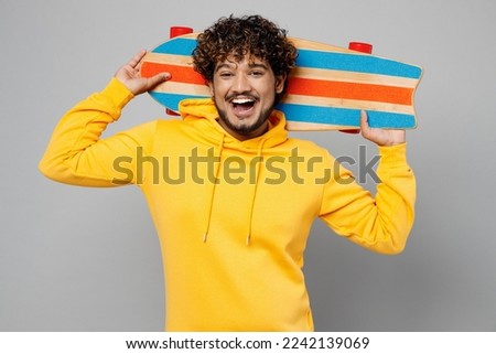 Young smiling happy fun cool cheerful Indian man 20s he wearing casual yellow hoody hold skateboard pennyboard look camera isolated on plain grey background studio portrait. People lifestyle portrait